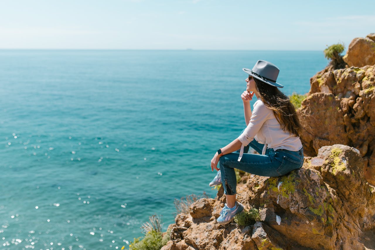 Woman in a hat sitting on a cliff enjoying the ocean view under a clear sky.