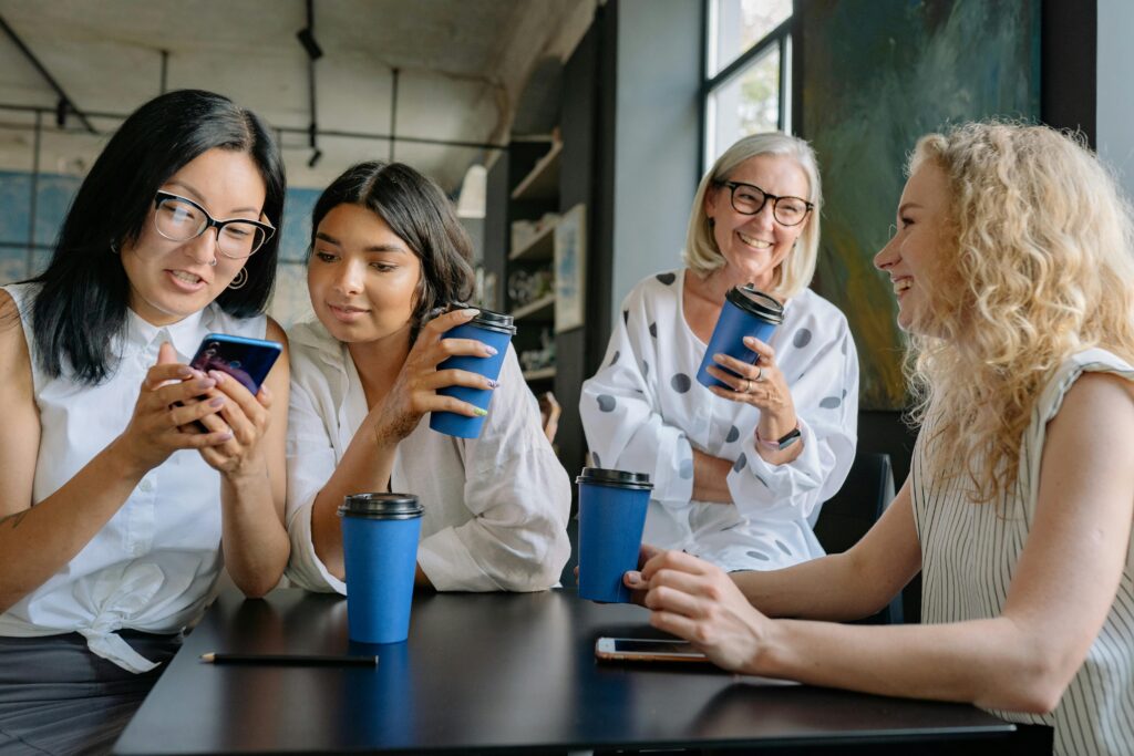 Four women of diverse ethnicities chatting and laughing during a casual coffee break in an office.